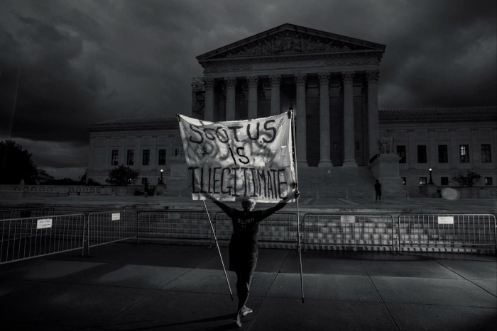 A black and white photo of a lone protester in front of an imposing classical portico holding a banner that reads ‘SCOTUS is illegitimate’