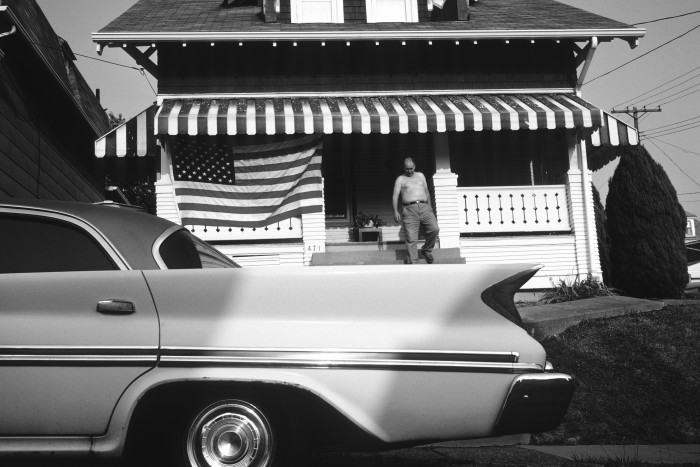 A black and white photograph of a big 1950s-style American car with pointed rear fins in front of a house displaying the US flag