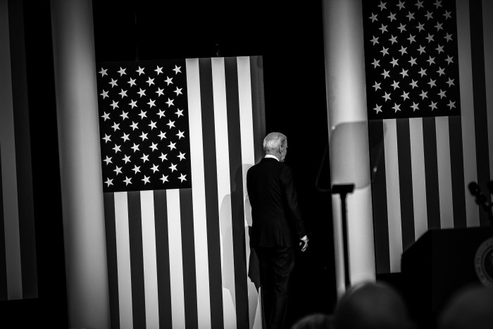 A black and white photo of Joe Biden, seen from behind, in front of a US flag
