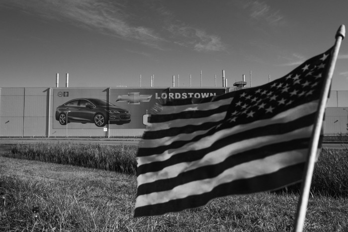 A stars and stripes flag flutters in the wind in front of a boarded up side with a photograph of a car