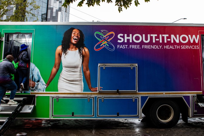 A large, brightly coloured mobile health clinic with “SHOUT-IT-NOW” branding is parked under a tree in a city area.