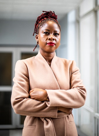 A strong and stylish woman with bold red lipstick and braided hair wears a beige coat while standing in a well-lit hallway