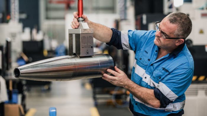 A man in a blue work uniform is carefully inspecting a large artillery shell in a factory setting.