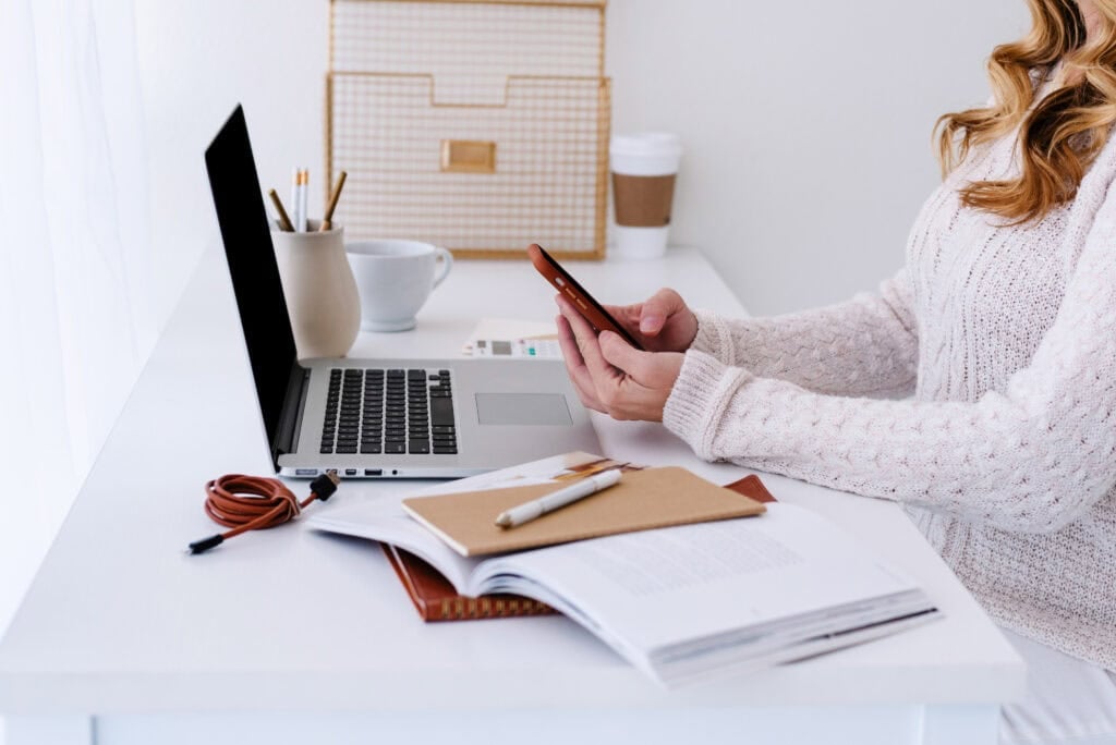 woman working on phone and laptop at desk - how to make money on fiverr for beginners