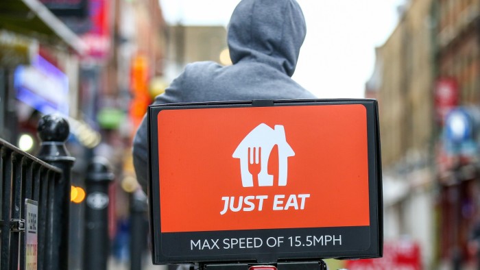 Back view of a food delivery courier wearing a gray hoodie riding a bicycle on Brick Lane in London. The delivery box on the back is branded with the "Just Eat" logo in white on an orange background, and it includes the text "MAX SPEED OF 15.5 MPH."