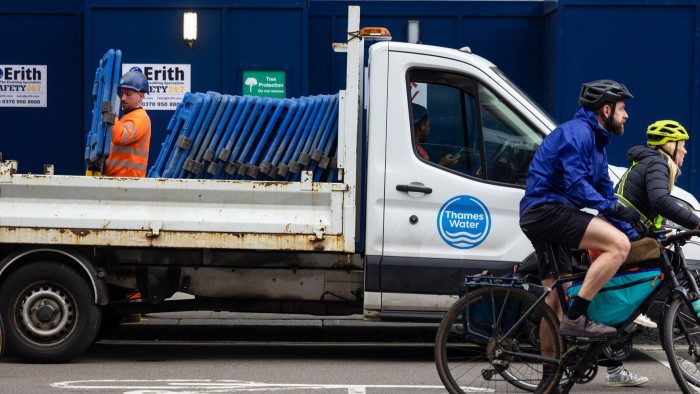 A Thames Water van is parked at a works location in London. An employee is removing protective barriers from the back of the van. Two cyclists ride past the van.