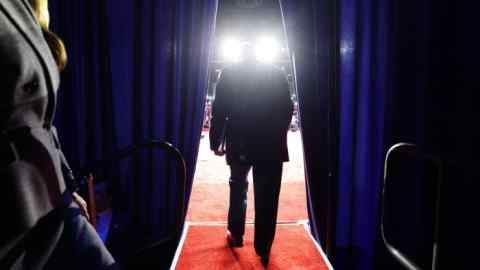 A bulky figure in a suit, seen from behind, walks through dark curtains on to a stage, a dazzle of lights partly obscuring his head