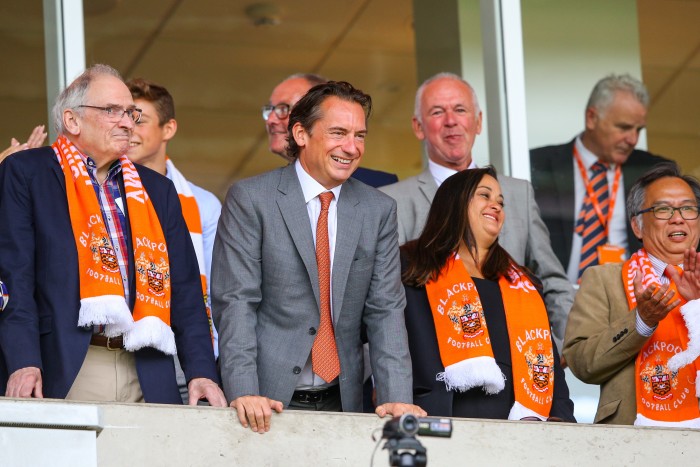 A group of seven people standing in a directors’ box looking out. Several wearing tangerine-coloured Blackpool Football Club scarves. In the centre is a man wearing a grey suit with a tangerine tie