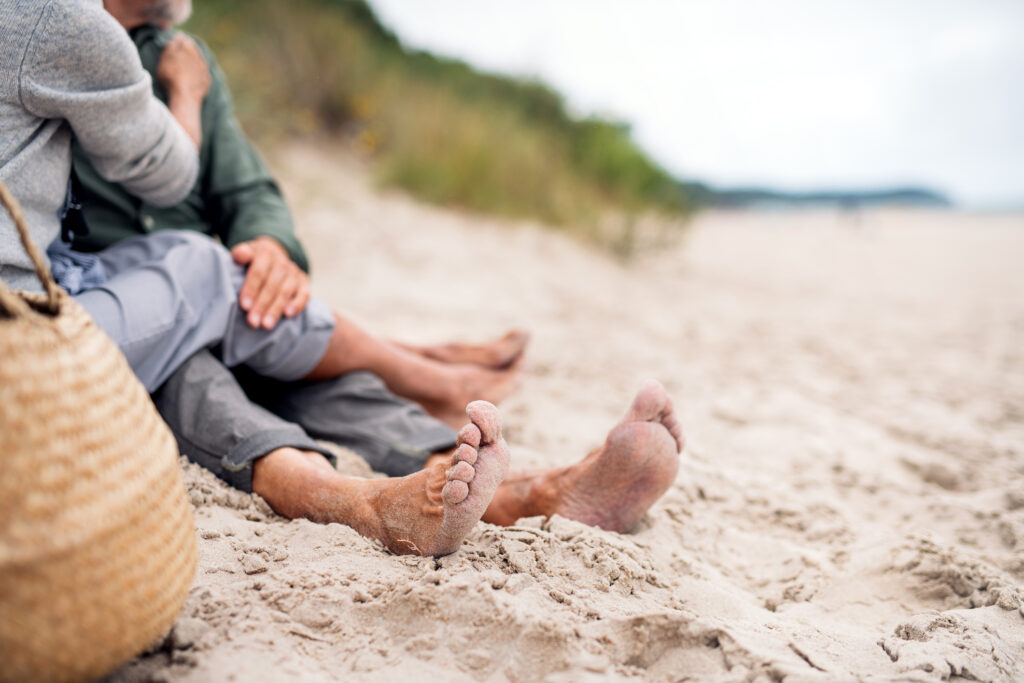 An older couple relaxes affectionately on a sandy beach with bare feet out. Their faces cannot be seen.