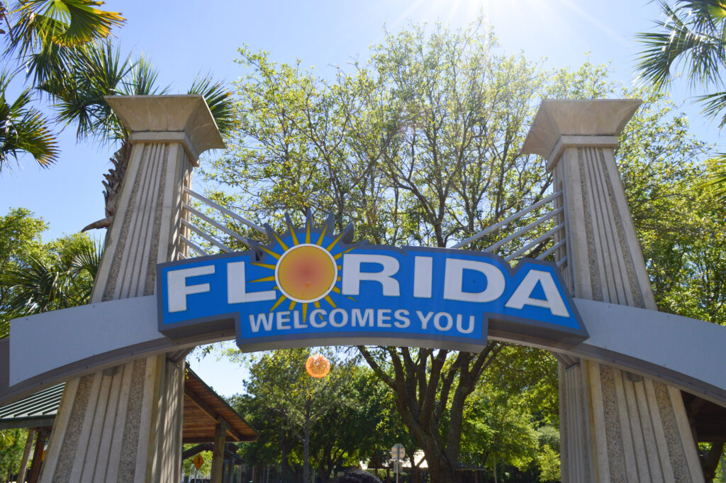 "Florida welcomes you" sign across two columns with trees in the background