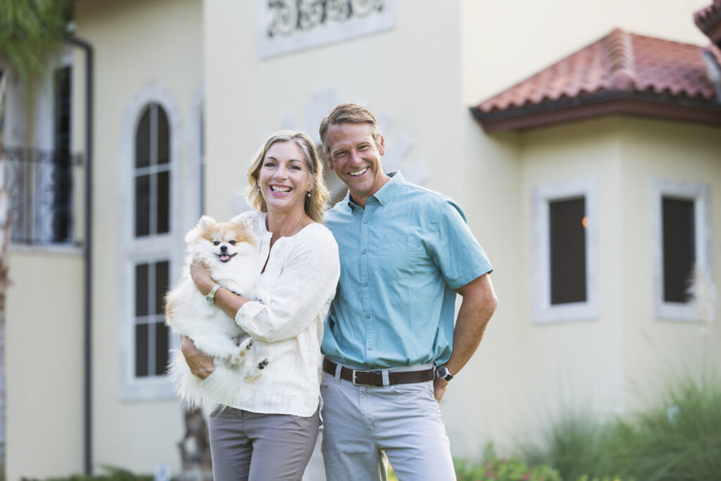 Portrait of an affluent mature couple standing in front of their home, a grand mansion. The woman is holding a cute pampered little dog in her arms while her husband stands beside her with his arm around her. They are smiling at the camera.