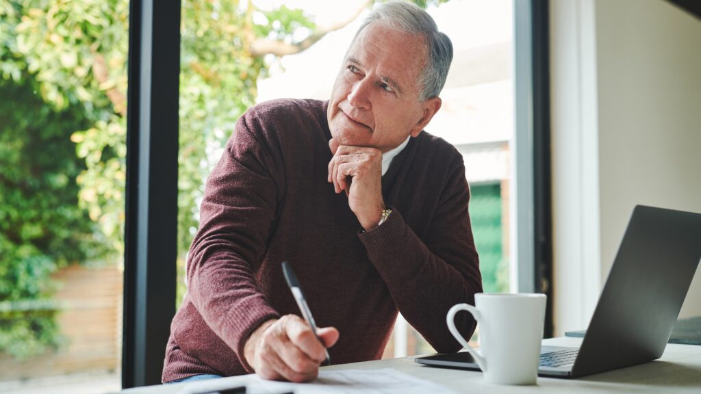 An older man thinks as he gazes into the distance while working on his laptop at a table.