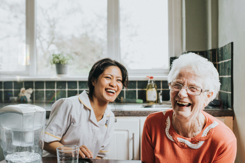 A young, friendly and bubbly carer sits beside an elderly woman at a kitchen table. The older woman faces the camera directly and laughs as the younger woman laughs behind her. The scene is lighthearted and fun.