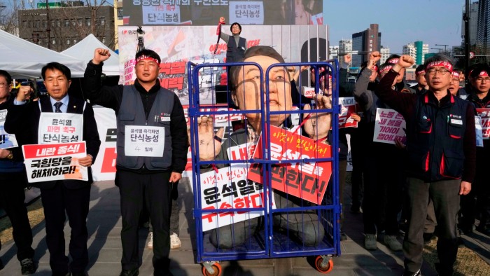 Protesters hold signs and raise their fists around a large effigy of impeached Yoon Suk Yeol enclosed in a cage, with signs calling for his immediate dismissal