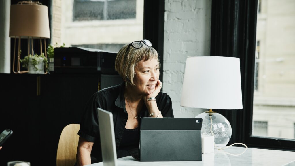 An older woman sits at her office desk, looking happily thoughtful.