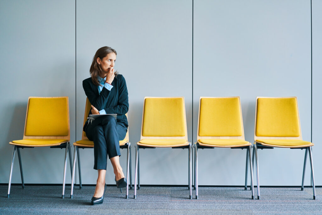 A woman in business clothes sits waiting in a room with yellow chairs.