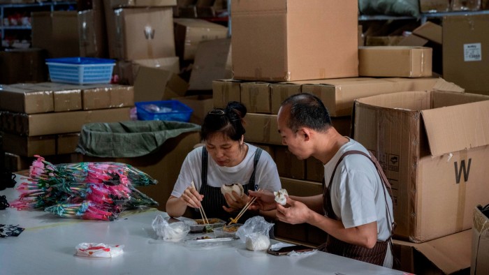 Workers at a factory sit together having lunch surrounded by stacked cardboard boxes and packaged festive goods