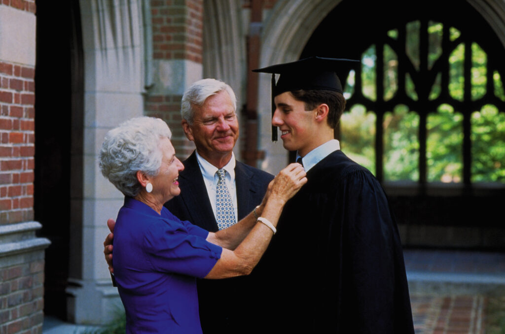 A grandson is graduation from college, talking with his grandparents. His grandmother is straightening his tie. They look close and happy.