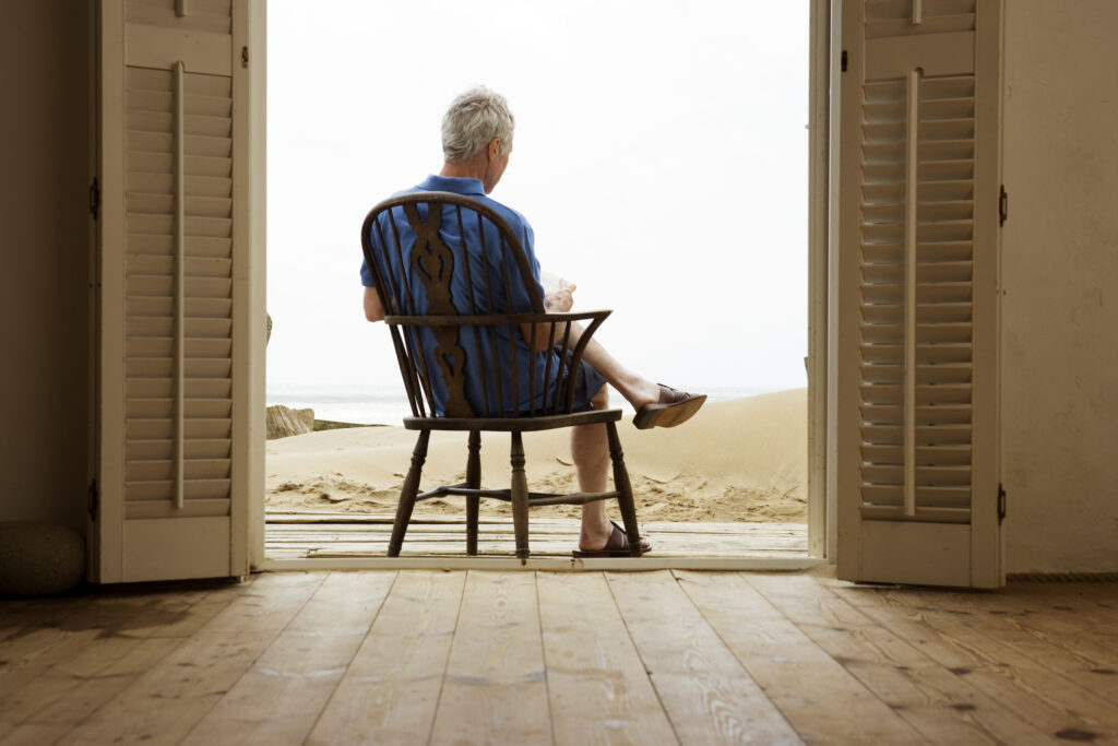An older man sits in a chair in the doorway to a beach house.