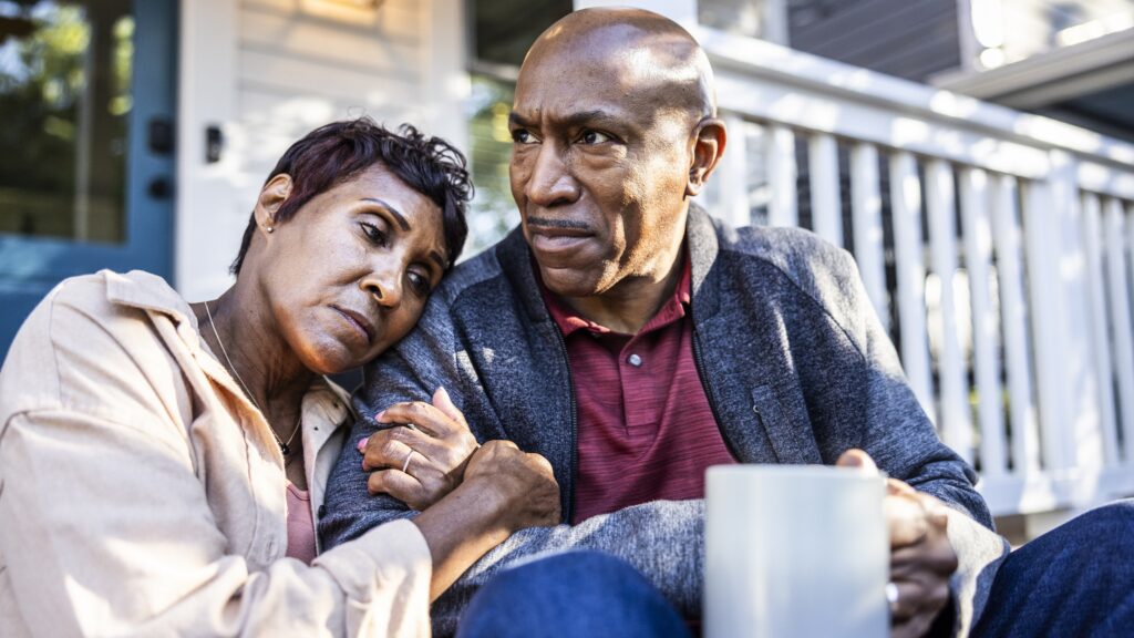 A retired couple sitting together on their front porch look stressed.