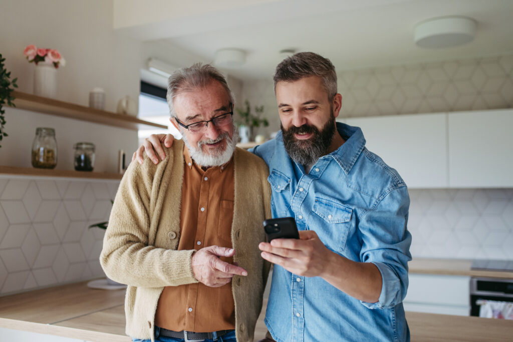 An adult son puts his arm around his father as they look at a phone together, standing in a kitchen.