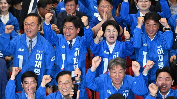 Members of the party of Korea are seen celebrating as they watch a television report on the exit poll. They are wearing blue jackets and expressing excitement with raised fists and smiles