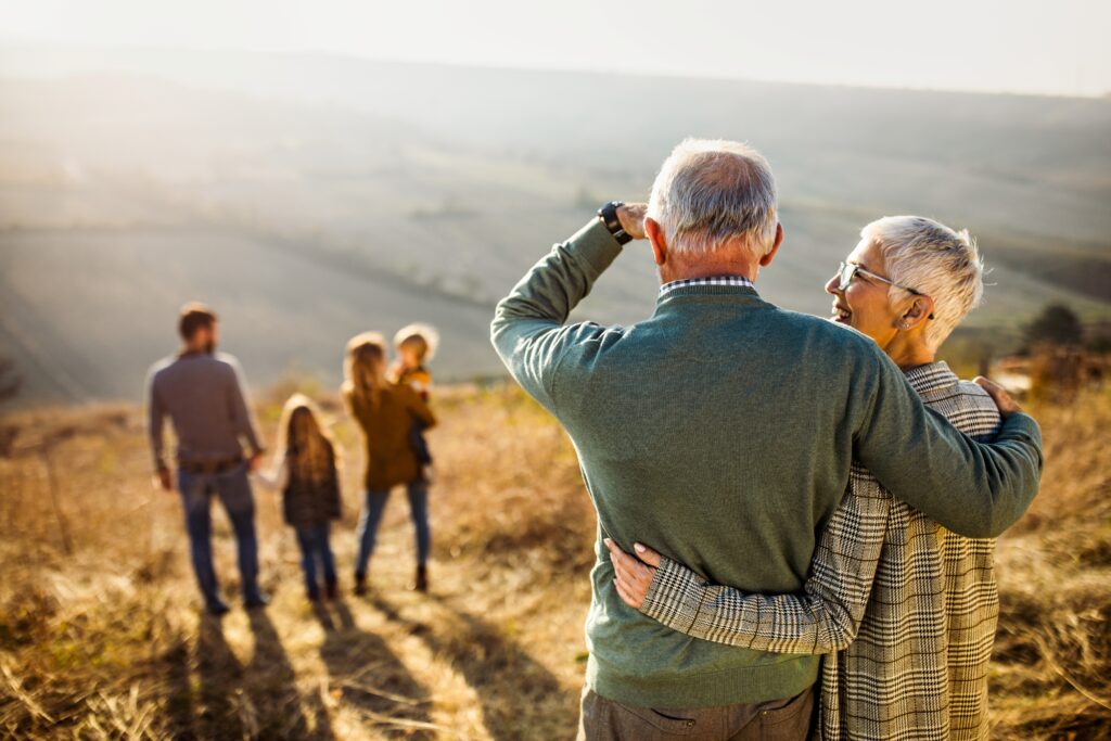 Grandparents stand together while smiling at their family in the distance.
