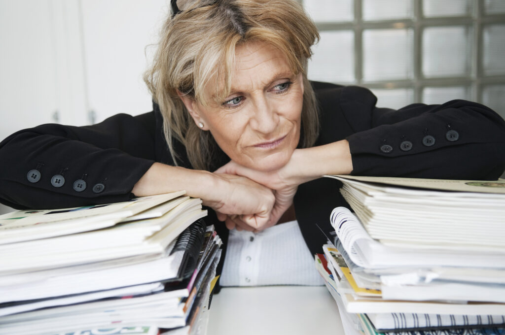 An older professional woman leans over two stacks of documents. She looks bored.