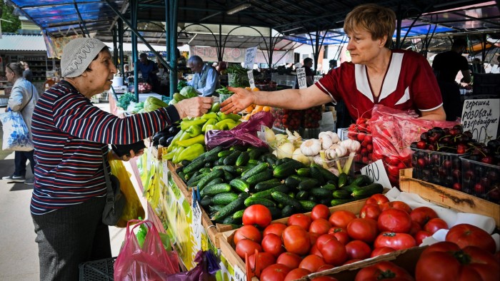A woman pays a vendor for green vegetables at an outdoor market in Moscow. The stall displays a variety of fresh produce