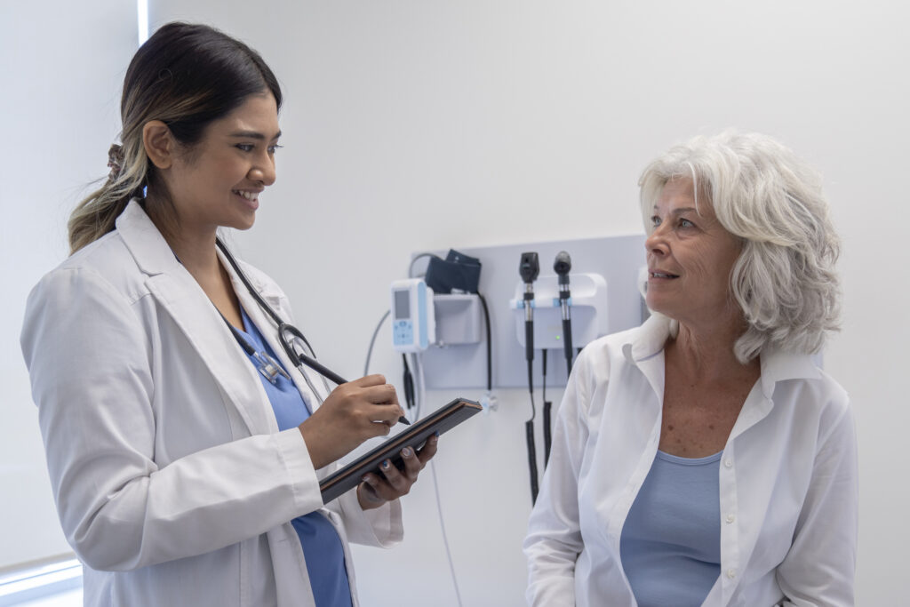A young female medical doctor is meeting with a senior female patient. She is smiling as she is looking at her patient.