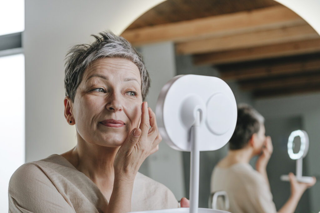 A woman with short grey hair applies moisturizer looking in a handheld mirror.