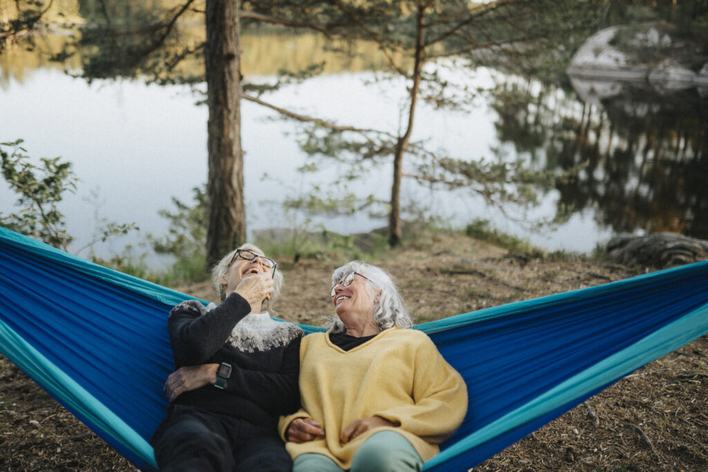 Two older women relax in a hammock by a lake, laughing.