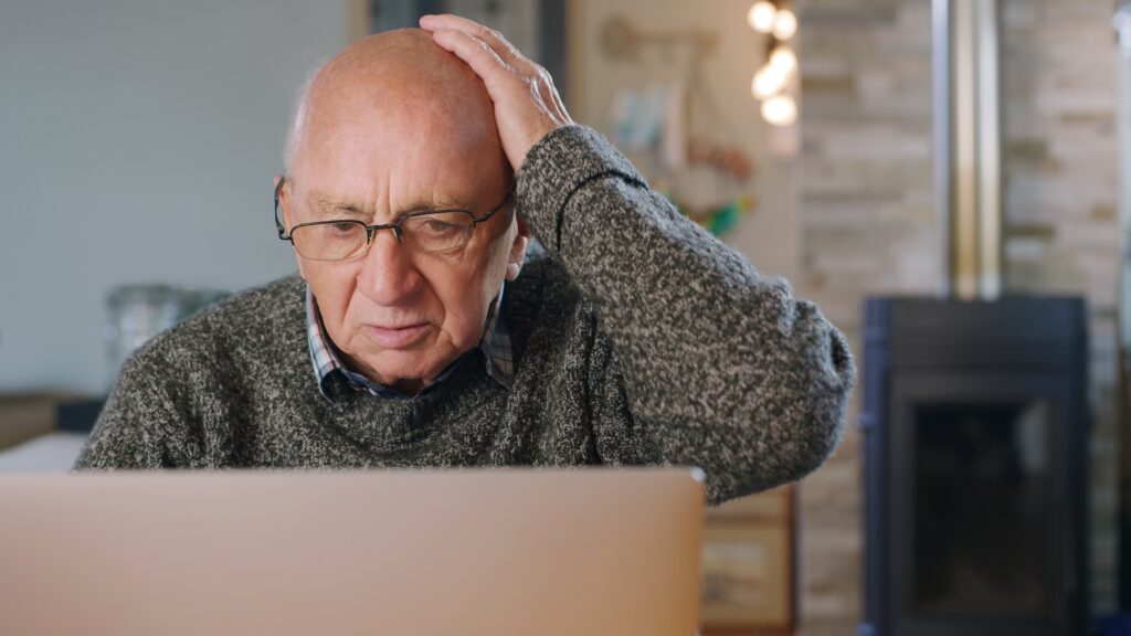 An older man looks confused or surprised while looking at his laptop at home.