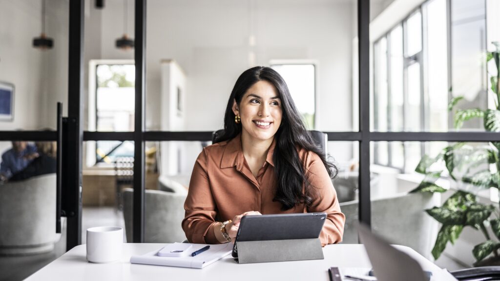 A businesswoman smiles as she sits at her desk in an office and looks into the distance as if thinking.