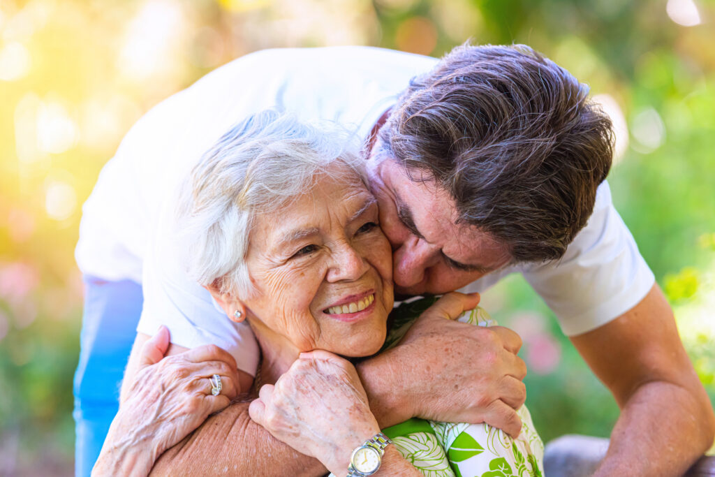 Mature man hugging his senior smiling mother while visiting her at a nursing home.