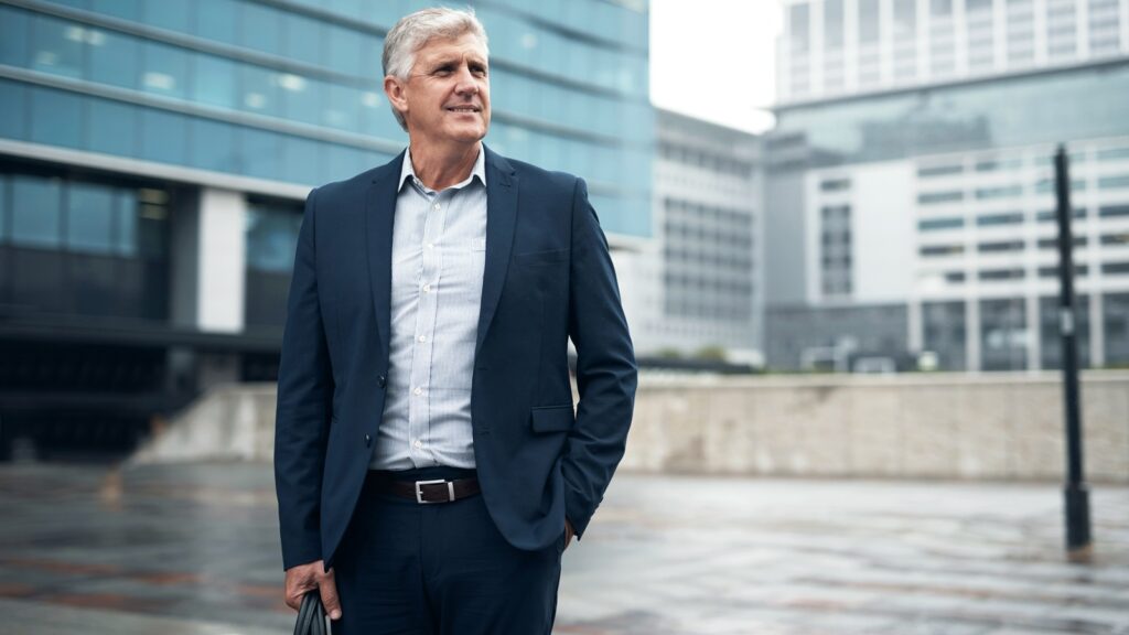 An older businessman stands outside his office.