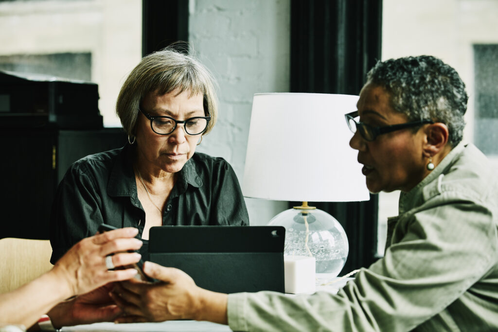 Two older women meet at a table, discussing something over a computer tablet.