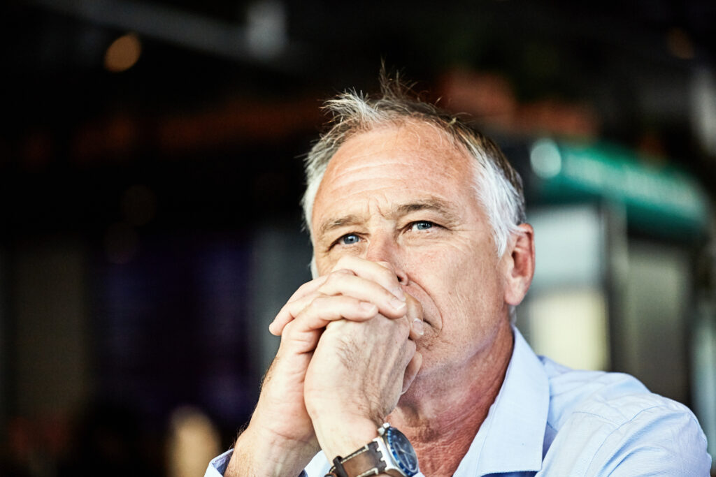 A retired man clasps his hands in front of his mouth and looks into the distance, behind the camera. He appears thoughtful or worried.