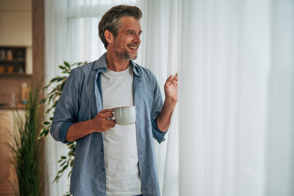 An older man smiles and looks out the window, holding a coffee cup.