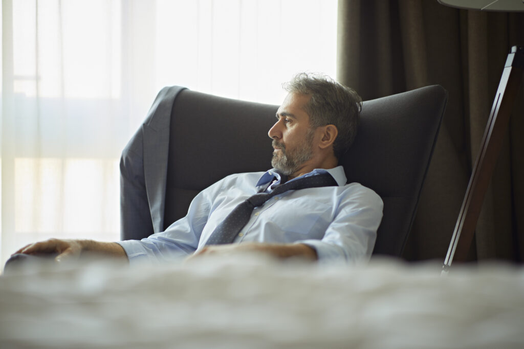 Businessman sitting in armchair in hotel room. His tie is loosened and he appears tired or burned out.
