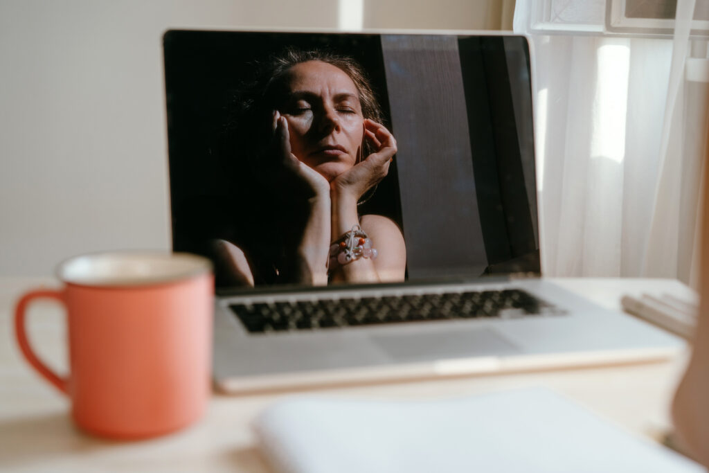 Portrait of a woman sitting at the notebook, red cup of tea on the table. Reflection of a face in the monitor screen, chin on hands. She looks tired.