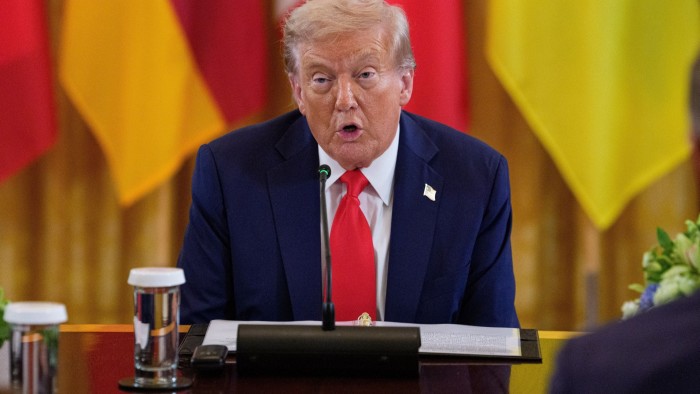 Donald Trump speaks at a table with a microphone, with blurred colourful flags in the background