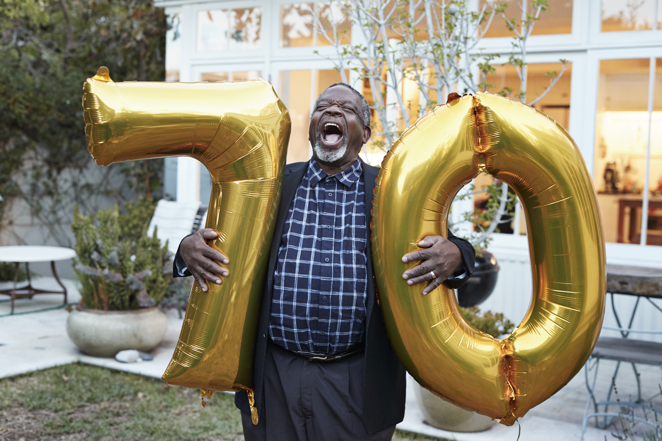 Senior man laughing while holding number 70 helium balloons in backyard during birthday party