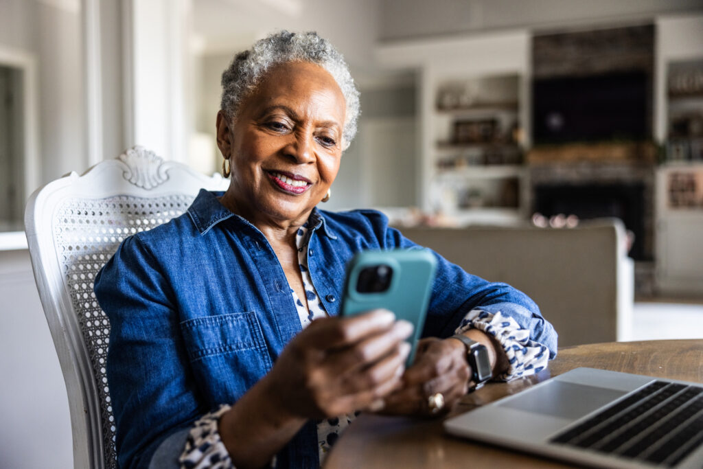 Older woman smiling and looking at cell phone while she sits at dining room table.