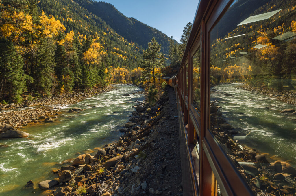A river and a mountain scene reflect in the window of a passenger railroad car going through an autumn scene. Photo taken October 15 2022 near Durango Colorado, USA.