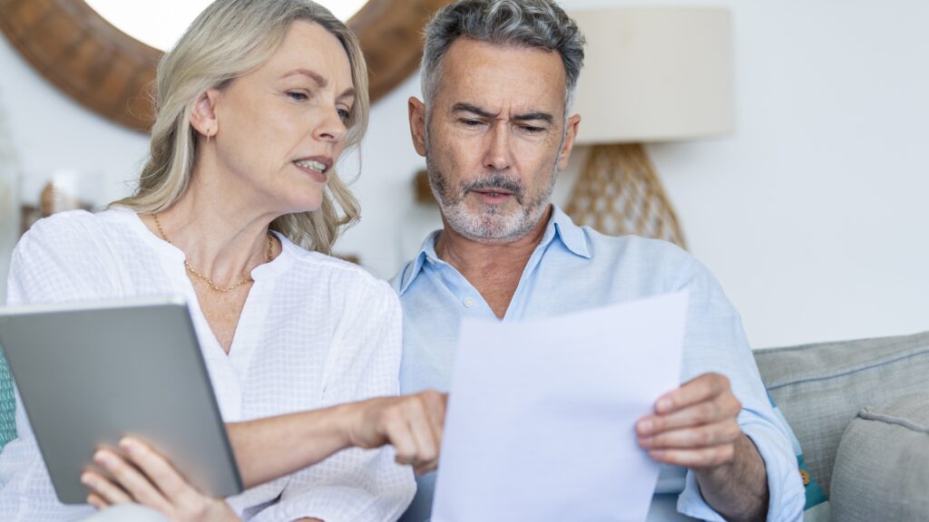 An older serious-looking couple look at a tablet together while sitting on their sofa.