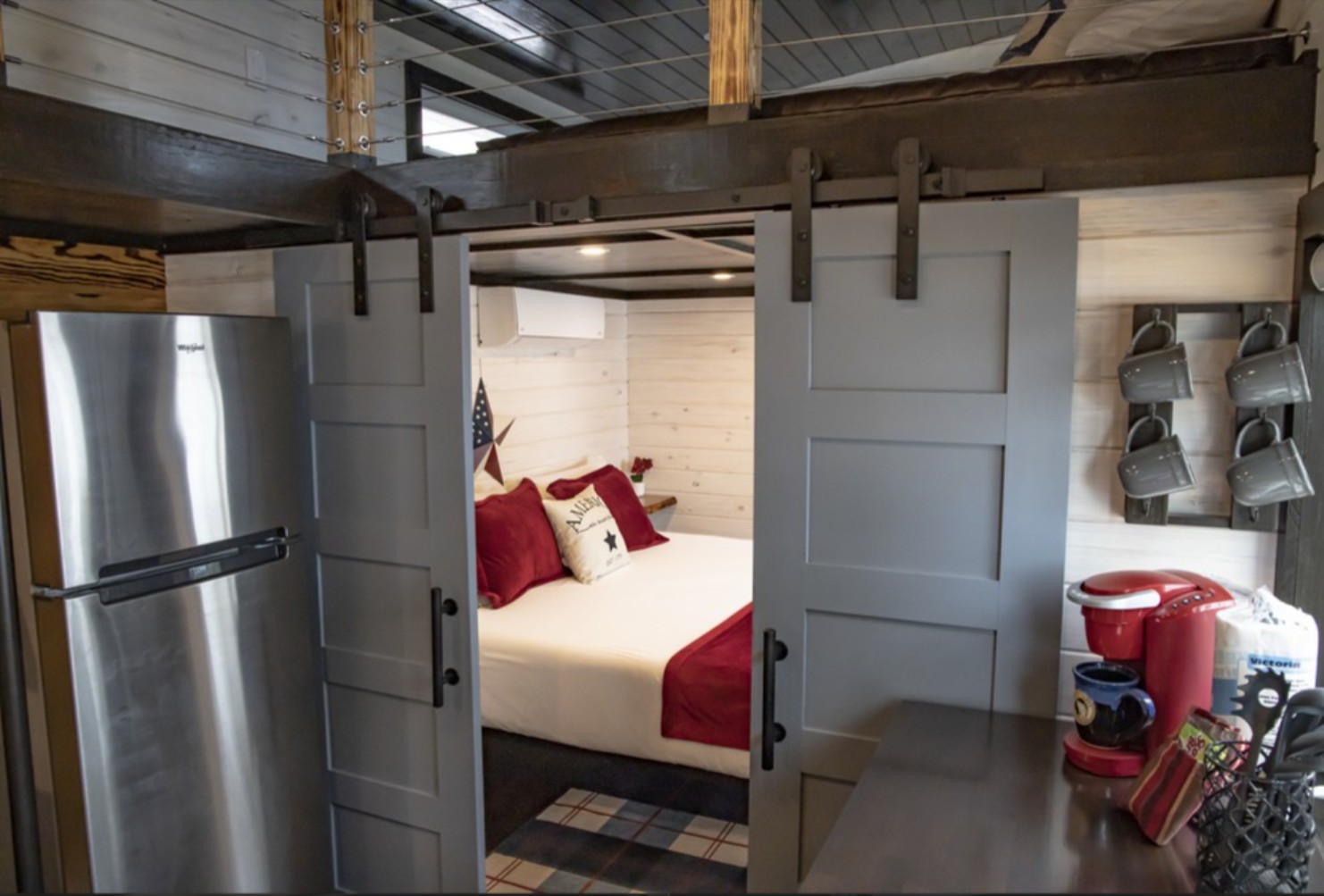 View of a comfortable, large bed in a tiny lakehouse (glamping cabin). The bed is visible through open farmhouse doors, with a small kitchen in the foreground. A sleeping loft is visible upstairs.
