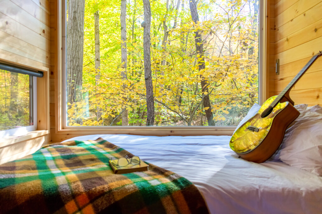 Interior of a glamping style tiny home in autumn in the woods. A guitar is propped on a comfortable looking bed by a picture window.