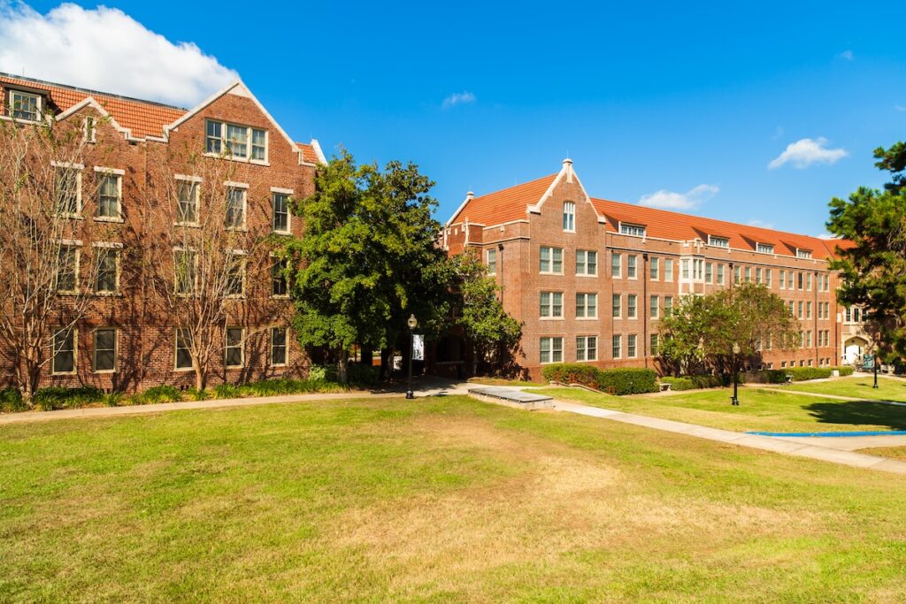 Sunny view of traditional red-brick college dormitories with clay-tiled roofs and manicured green lawns under a blue sky. This image visualizes the costly on-campus housing facilities that make up the