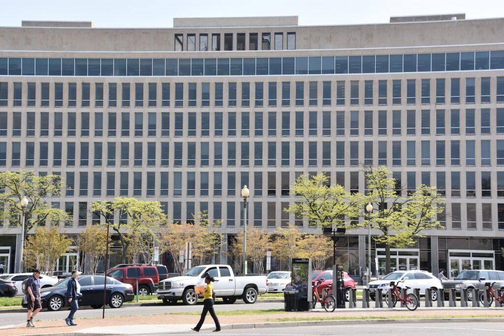 Wide-angle view of the U.S. Department of Education headquarters in Washington, D.C., featuring its distinct beige, grid-patterned facade towering over a busy street with pedestrians and vehicles. This government building serves as the focal point for this week's major news, specifically the department's controversial move to transfer six education programs to other federal agencies. | Photo by sainaniritu